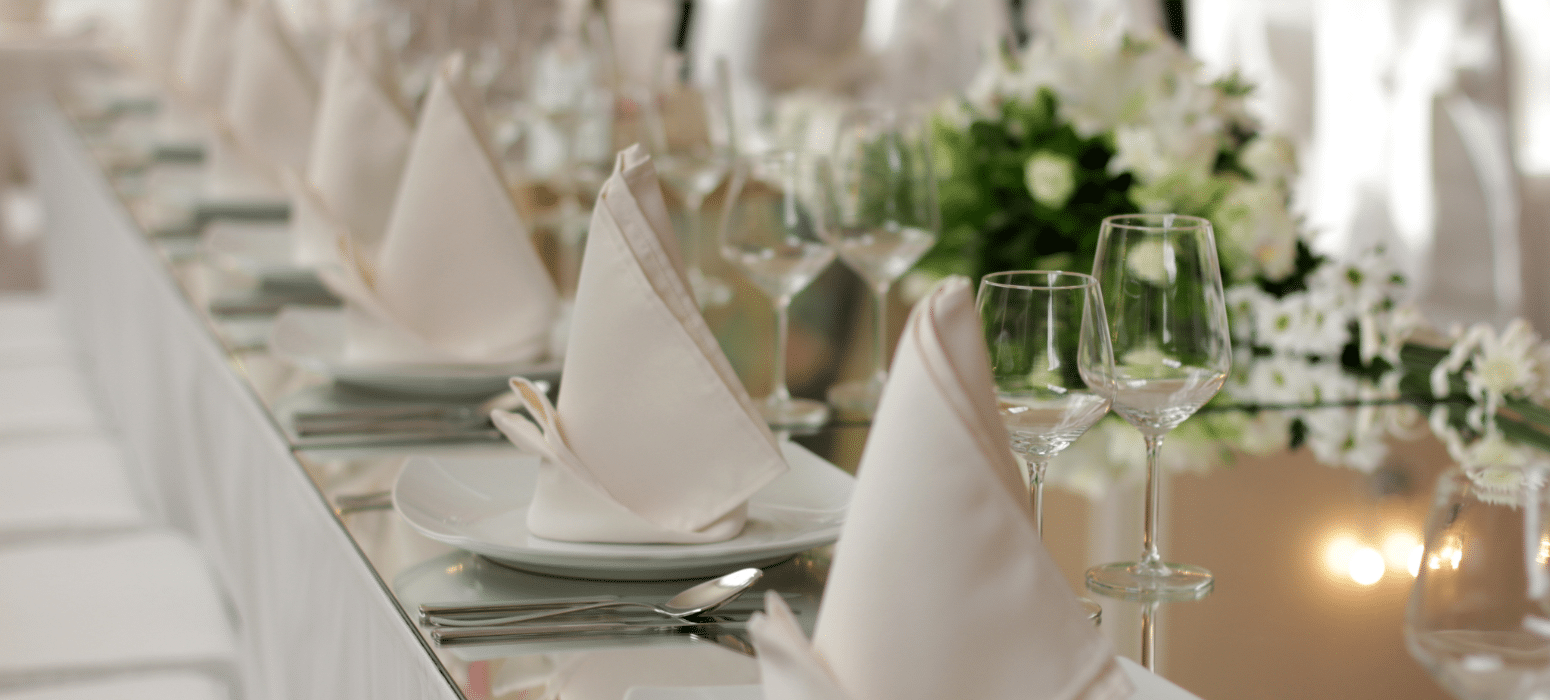 A close-up of an elegant table setting with white napkins folded into triangles, white plates, silver cutlery, wine glasses, and a floral centerpiece in the background.