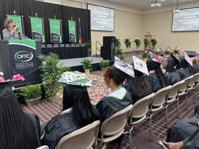 Graduates in caps and gowns sit facing a stage at Oconee Fall Line Technical College’s commencement ceremony. A woman speaks at the podium while decorated graduation caps are visible among the seated graduates.