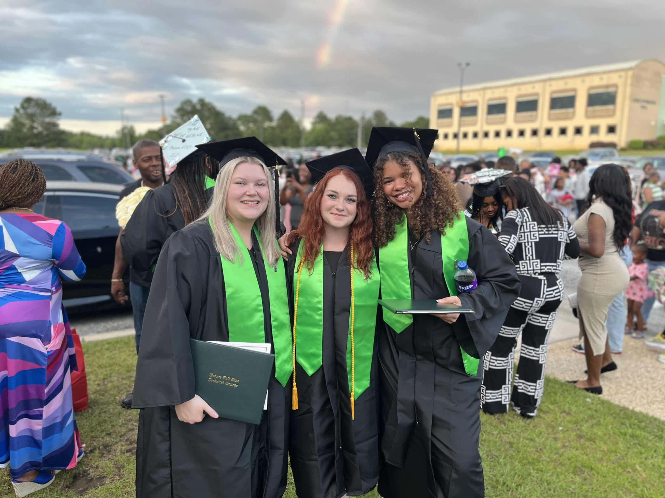 Three graduates in black caps and gowns with green stoles smile and pose together outdoors after a ceremony, holding diplomas. A rainbow is visible in the cloudy sky, and a crowd is gathered in the background.