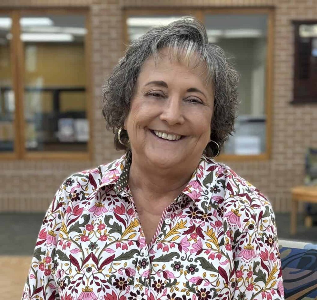 A smiling older woman with short gray hair wears a floral-patterned shirt and hoop earrings, standing indoors in front of large windows and a brick wall.