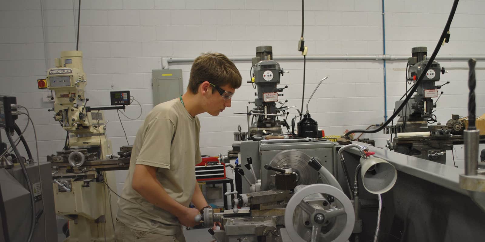 A person wearing safety glasses operates a metal lathe in a workshop, surrounded by various milling machines and industrial equipment.