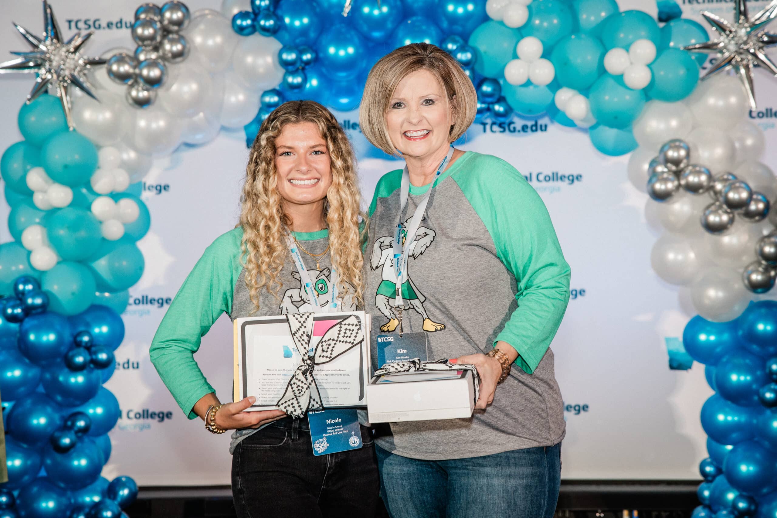 Two women wearing matching blue and green shirts stand smiling in front of a blue and white balloon arch, each holding gift boxes. The backdrop features the TCSG.edu logo.