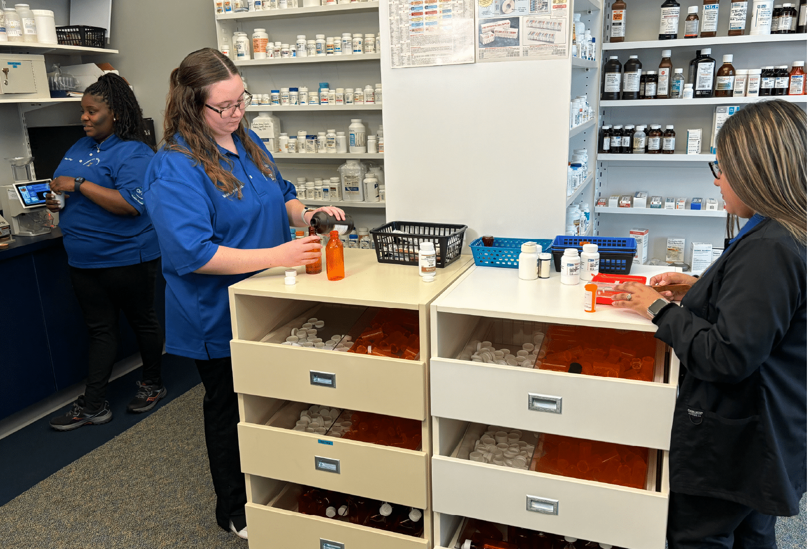 Three pharmacy employees, two in blue uniforms and one in black, prepare and organize medication bottles at a counter with drawers filled with empty pill bottles. Shelves with various medications are in the background.