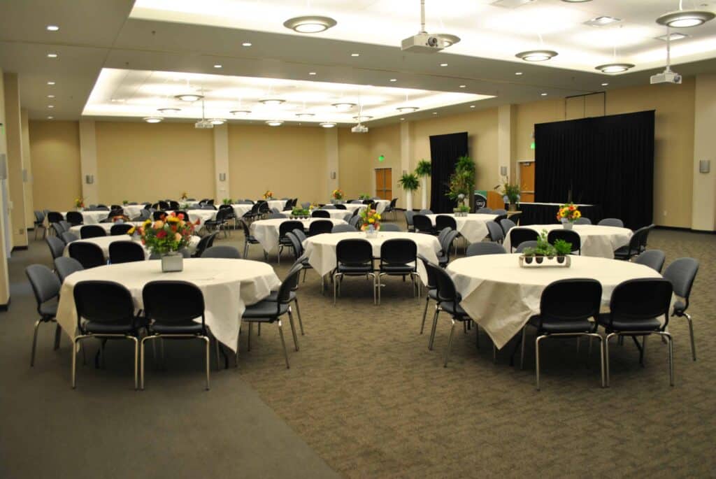 A large conference room with round tables covered in white tablecloths, each surrounded by chairs and decorated with flower centerpieces, set up for an event or banquet.