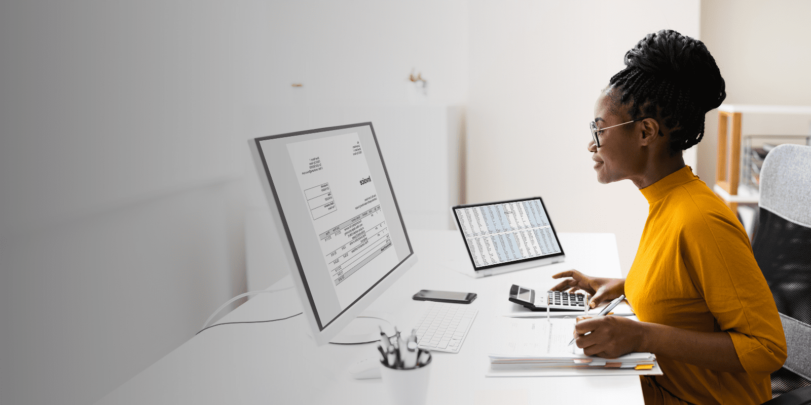 A woman wearing glasses and a yellow top sits at a desk, working on a computer with a financial document on the screen. She uses a calculator and has a notebook, keyboard, and smartphone nearby.