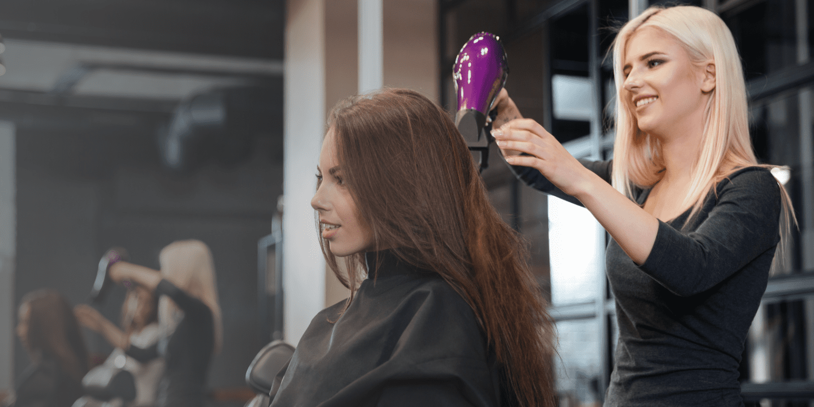 A smiling hairstylist blow-dries a young woman’s long brown hair in a modern salon. The client wears a black cape, and another stylist works with a customer in the background.