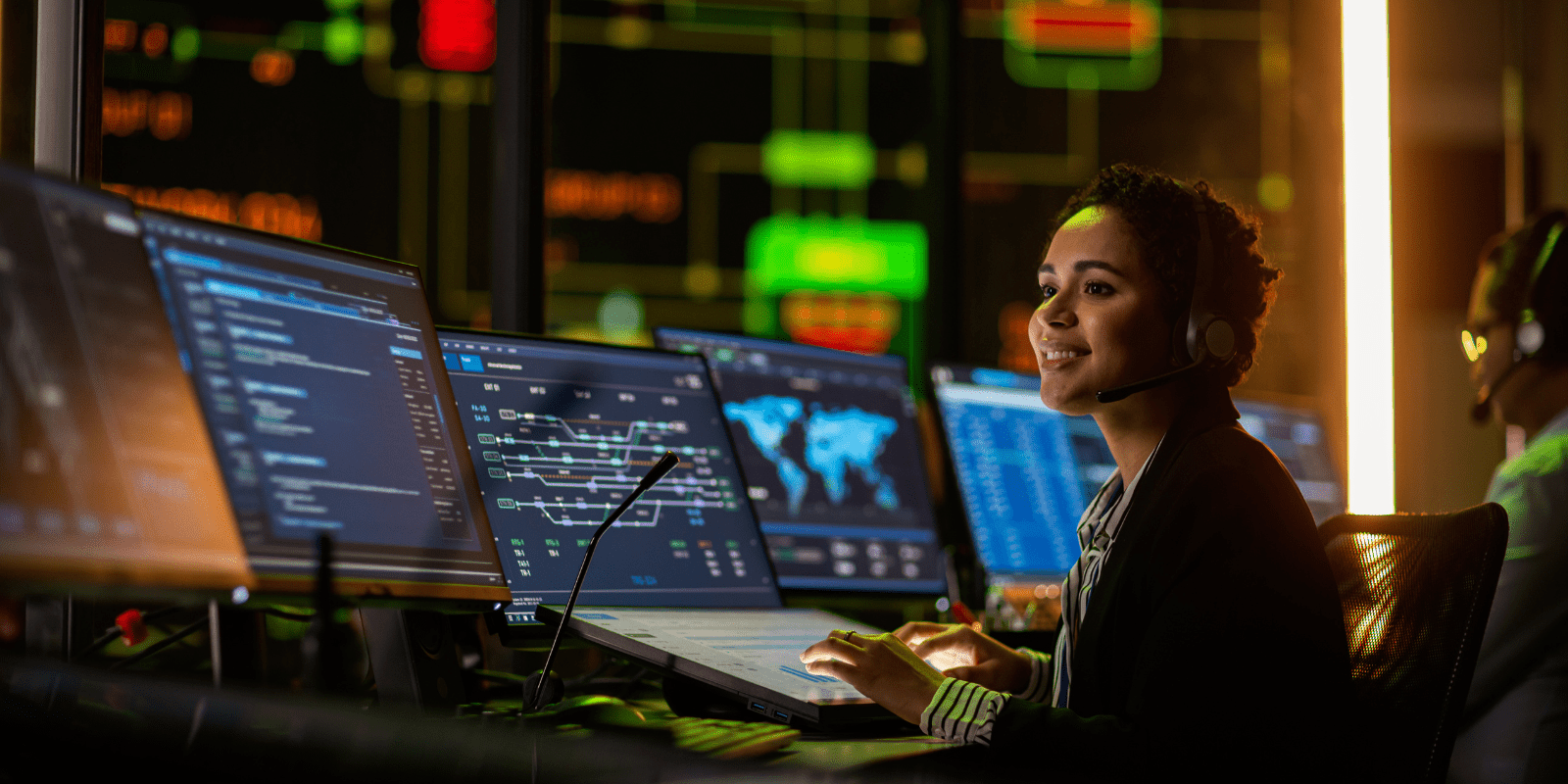 A woman wearing a headset sits at a control station with multiple computer monitors displaying data, maps, and digital systems in a high-tech operations center.