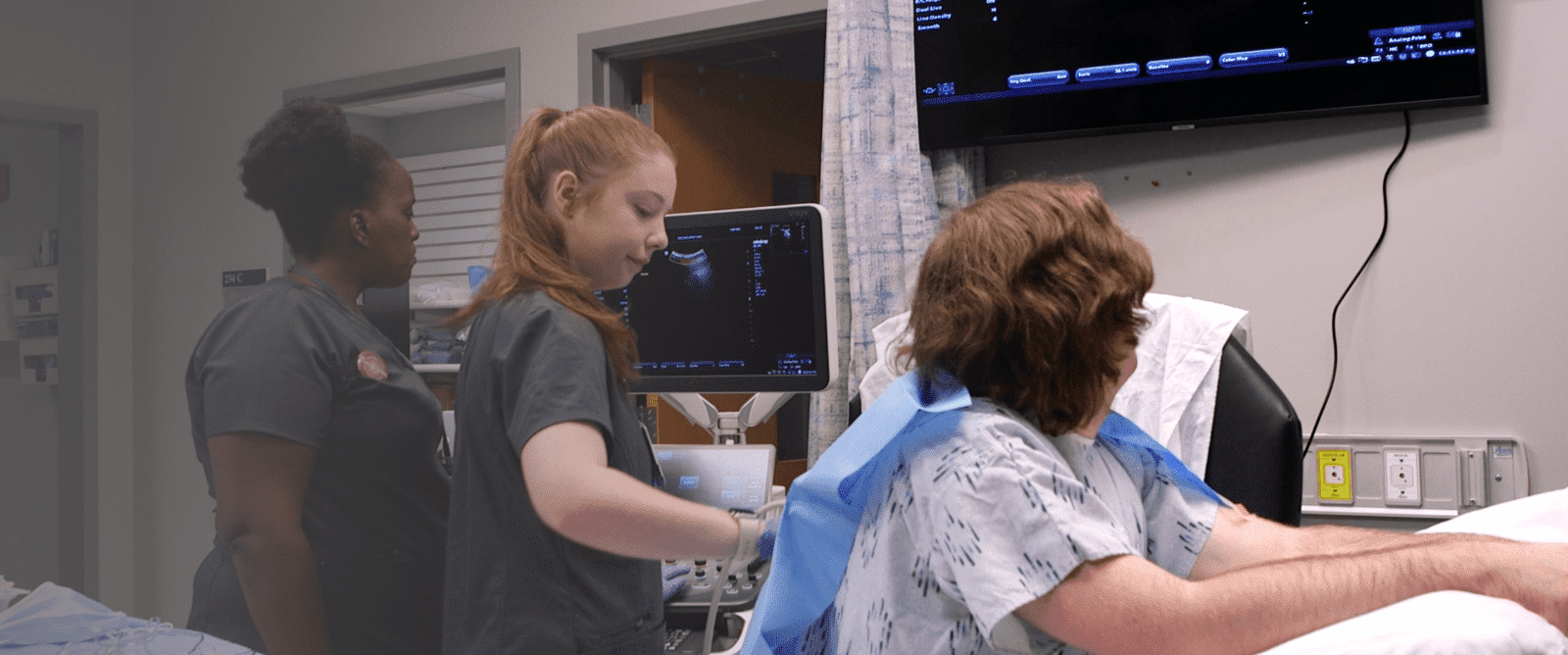 Two medical professionals in scrubs perform an ultrasound on a patient lying on a bed with medical monitors and equipment visible in a clinical setting.
