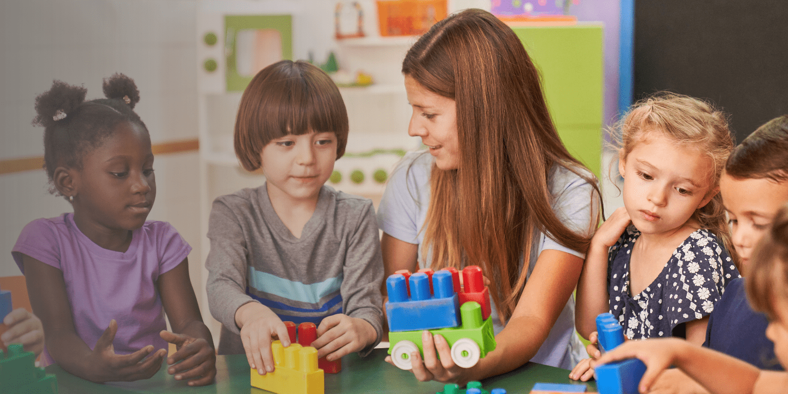 A teacher sits with a group of young children at a table, playing and building with colorful plastic blocks in a classroom setting.