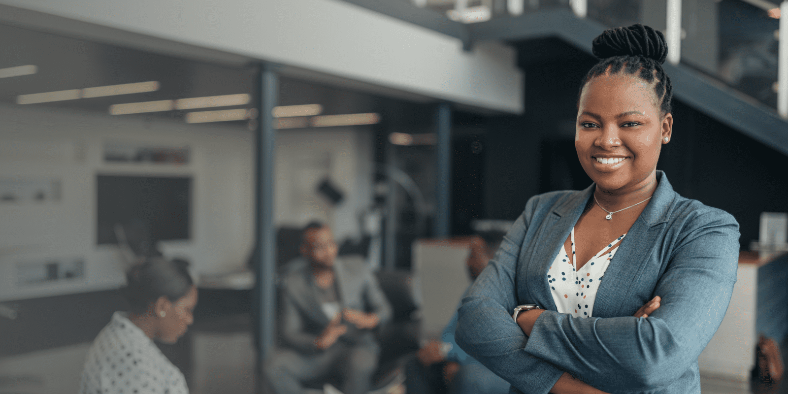 A confident woman in a business suit stands with arms crossed and smiling in an office, while colleagues have a meeting in the blurred background.