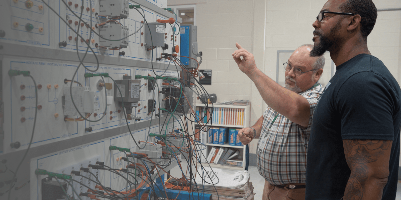 Two men stand in front of an electrical control panel with wires and equipment; one man points at the panel while explaining something to the other in a technical classroom or lab setting.
