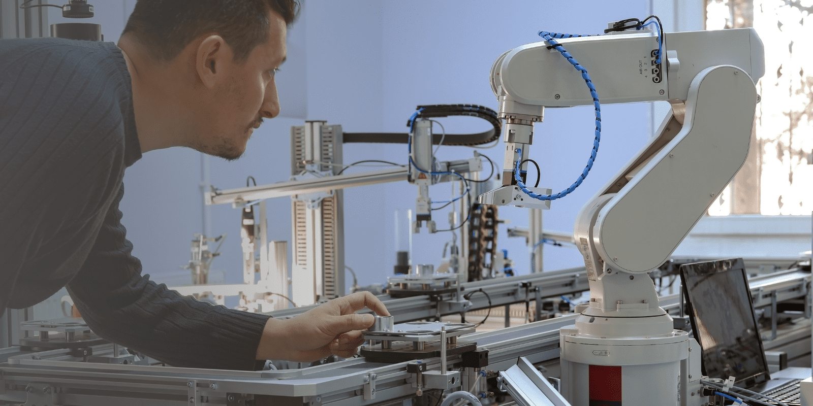 A man interacts with a robotic arm in a lab setting, adjusting equipment on a workbench surrounded by various machines and technology components.