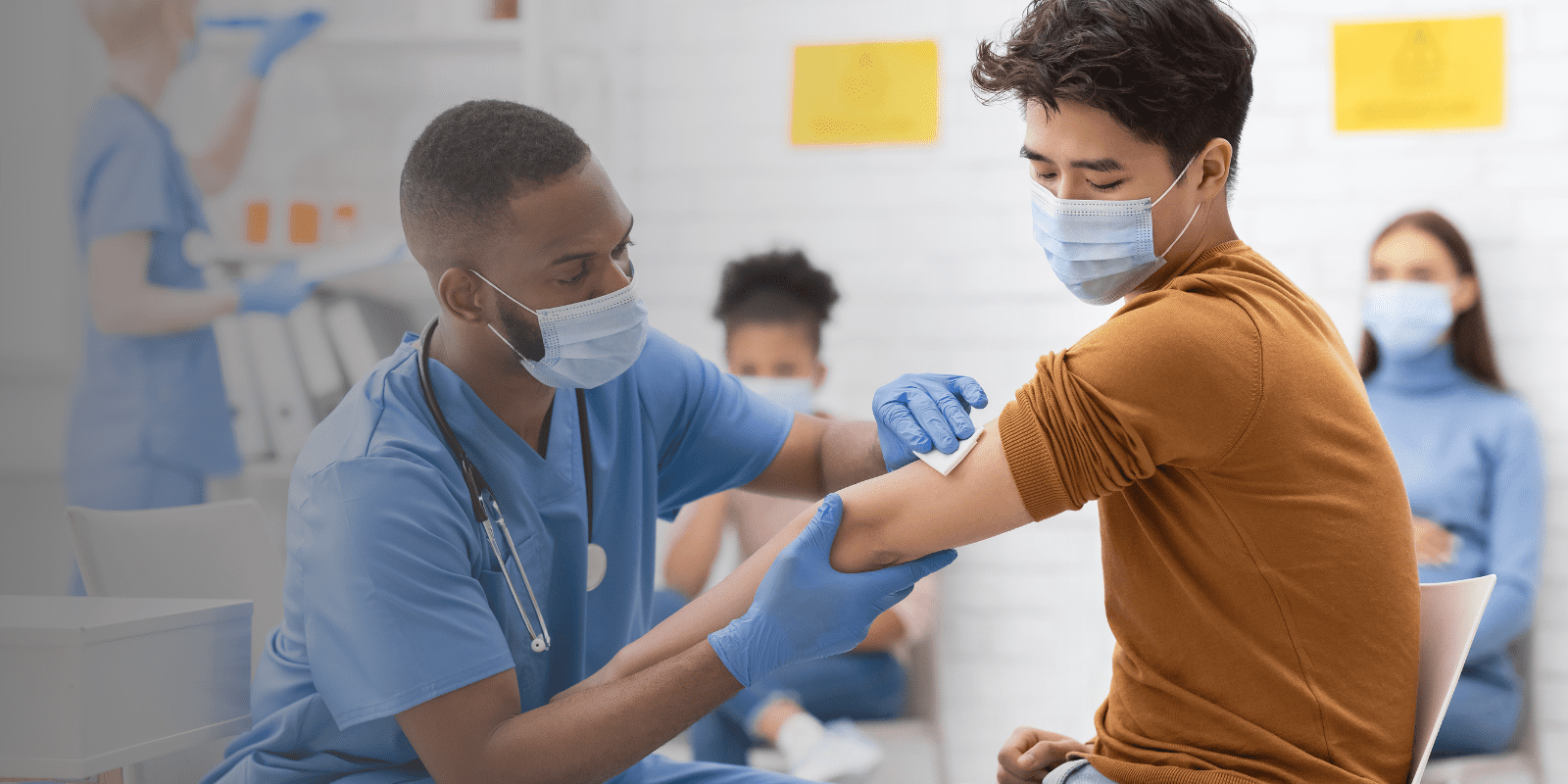 A healthcare worker in scrubs prepares to give a vaccine to a young man wearing a mask, while other masked individuals wait in the background.