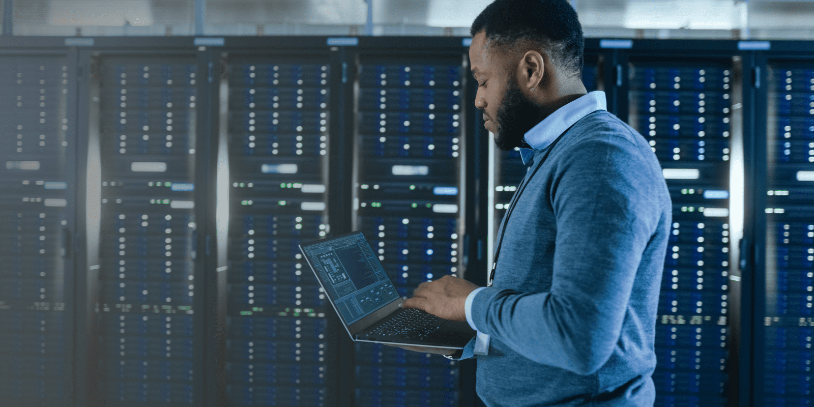 A man stands in a server room working on a laptop. Behind him are several tall server racks filled with blinking lights and electronic equipment. The setting suggests a data center or IT environment.