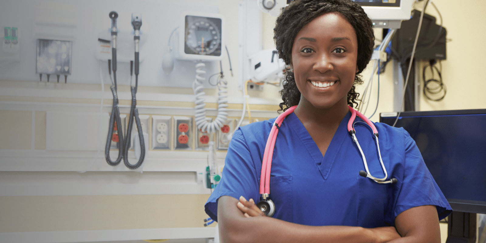 A smiling healthcare professional in blue scrubs with a stethoscope around her neck stands confidently with arms crossed in a modern medical examination room.