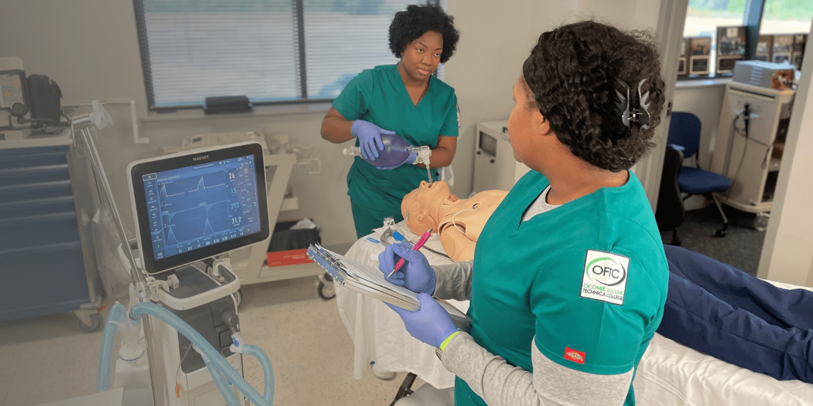 Two healthcare students in green scrubs practice medical procedures on a mannequin in a clinical skills lab, one recording notes and the other using a manual resuscitator. Medical equipment and monitors are visible nearby.