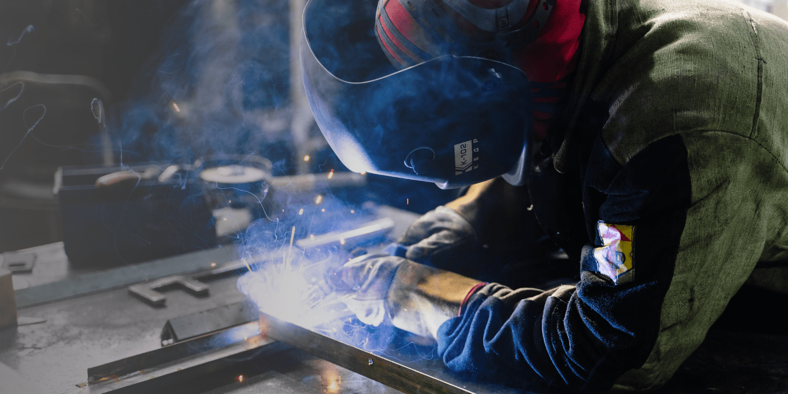 A person wearing protective gear and a welding mask welds metal, producing bright sparks and smoke in a workshop.