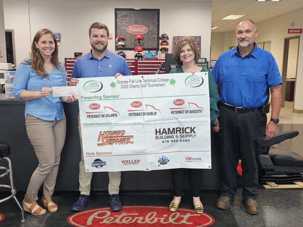 Four people stand indoors holding a large event sponsor sign and displaying a check. The sign lists sponsors for Oconee Fall Line Technical College 2025 Charity Golf Tournament. A Peterbilt logo is visible in the background.