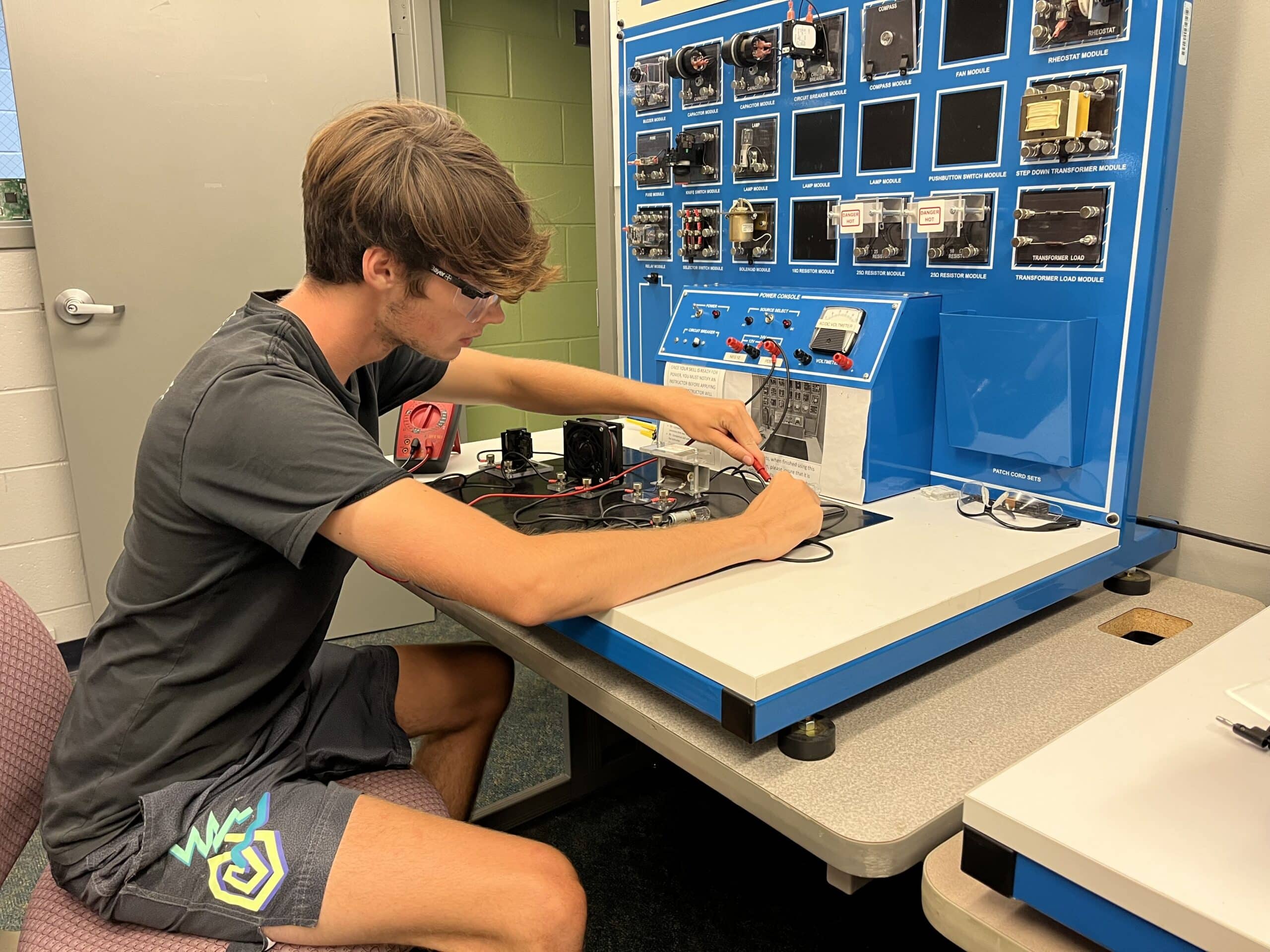 A young man wearing glasses and shorts sits at a desk working with electronic components and wires on a training board in a classroom or lab setting.