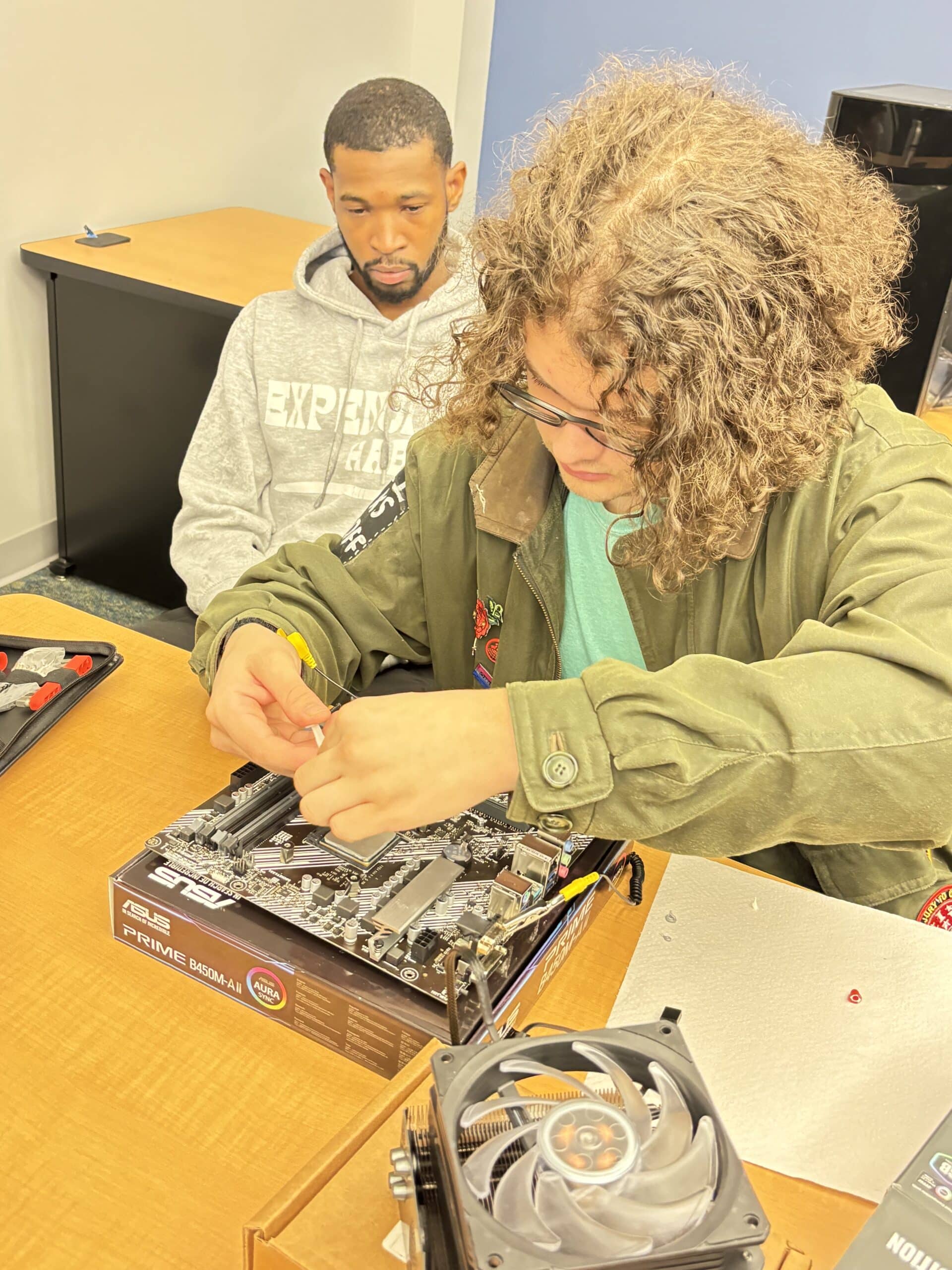 Two people sit at a table assembling a computer. The person in front, with curly hair and glasses, works on a motherboard, while the person behind watches attentively. Computer parts and tools are on the table.