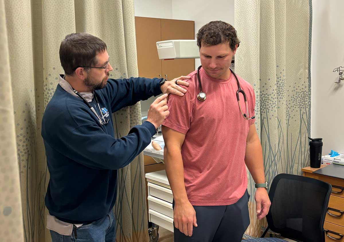 A healthcare professional examines a man's shoulder in a medical office. The man in a red shirt stands while the provider, wearing a stethoscope and blue jacket, gently checks his arm. Medical equipment is visible in the background.