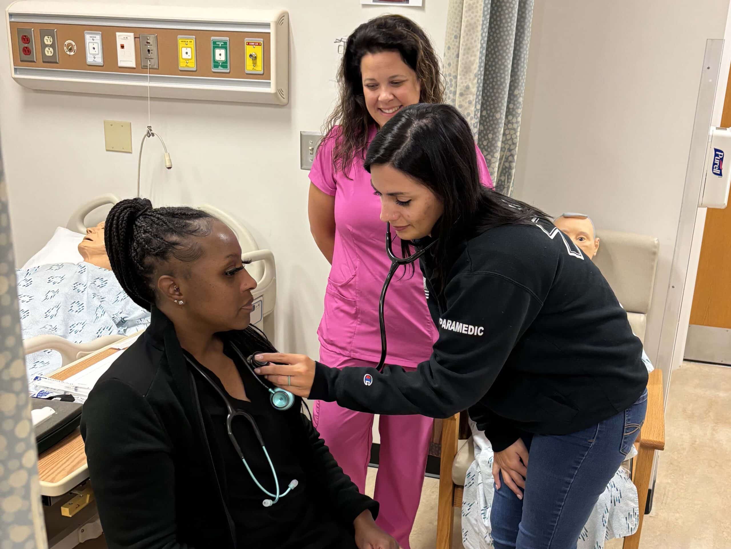 Three women are in a medical training room. One woman sits while another uses a stethoscope on her chest. A third woman in pink scrubs stands behind them, smiling. Medical beds and equipment are visible in the background.