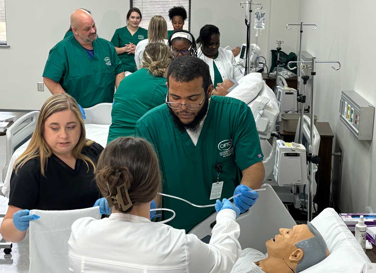 A group of medical students in green uniforms practice patient care and medical procedures on a mannequin in a clinical training room, guided by instructors in white coats.