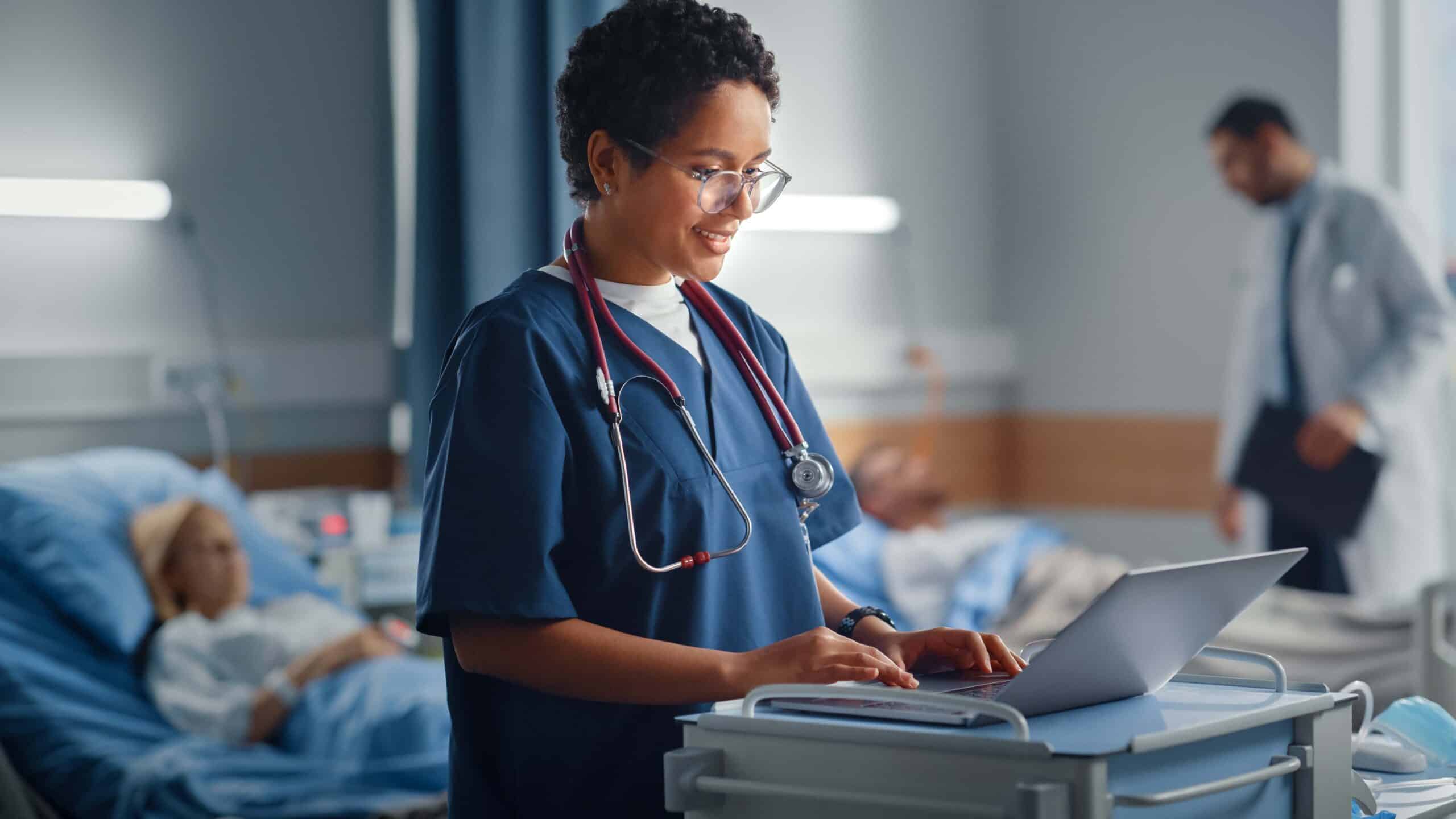 A smiling healthcare worker in scrubs and a stethoscope uses a laptop on a medical cart in a hospital room, with patients resting in beds and another medical professional in the background.