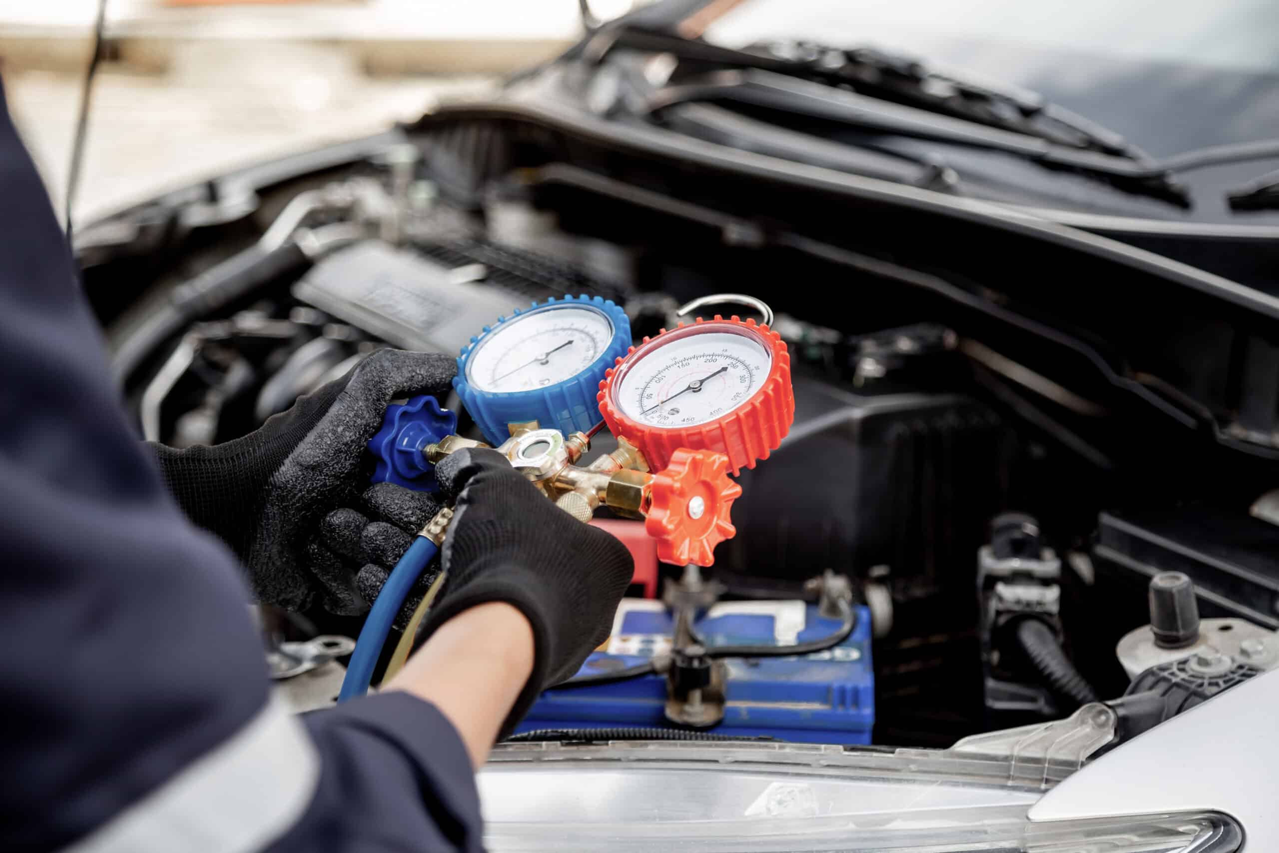 A person wearing black gloves uses a manifold gauge to check the air conditioning system under the hood of a car.