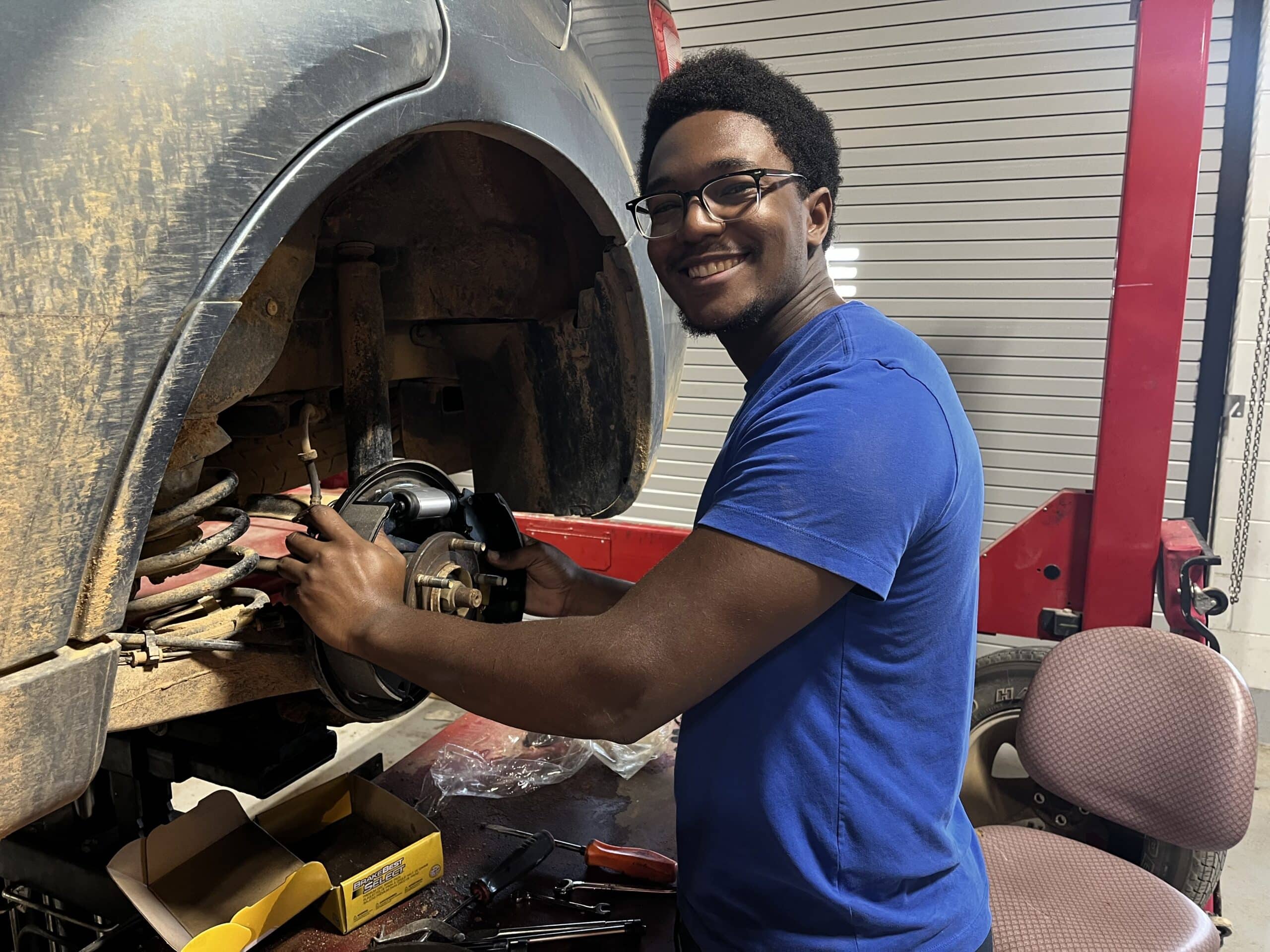 A person in a blue shirt smiles as they repair a car's brake system in a garage, with tools and parts nearby and the wheel removed.