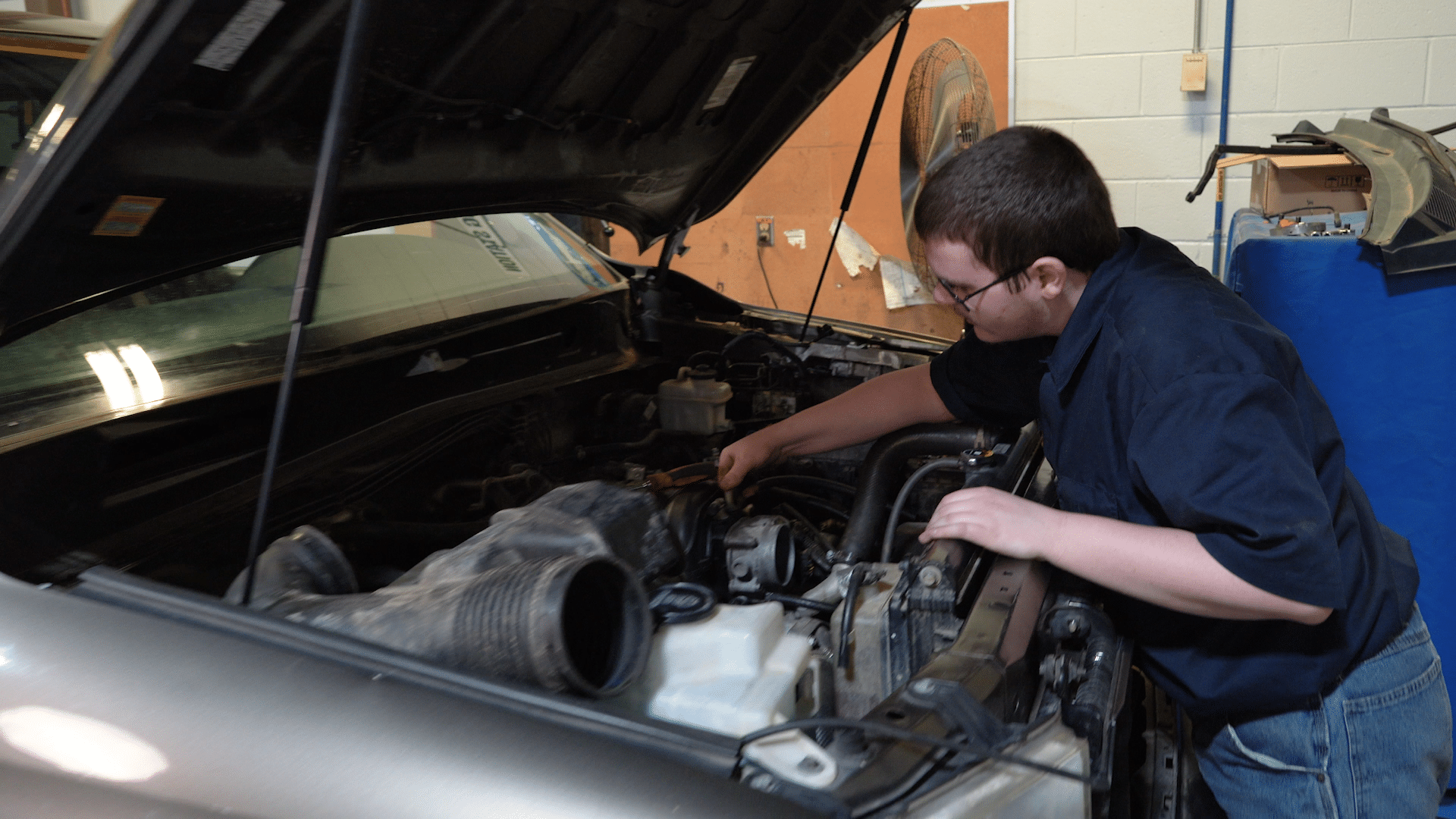 A person wearing glasses and a dark shirt works under the open hood of a car in a garage or workshop, using a tool to adjust or repair the engine.