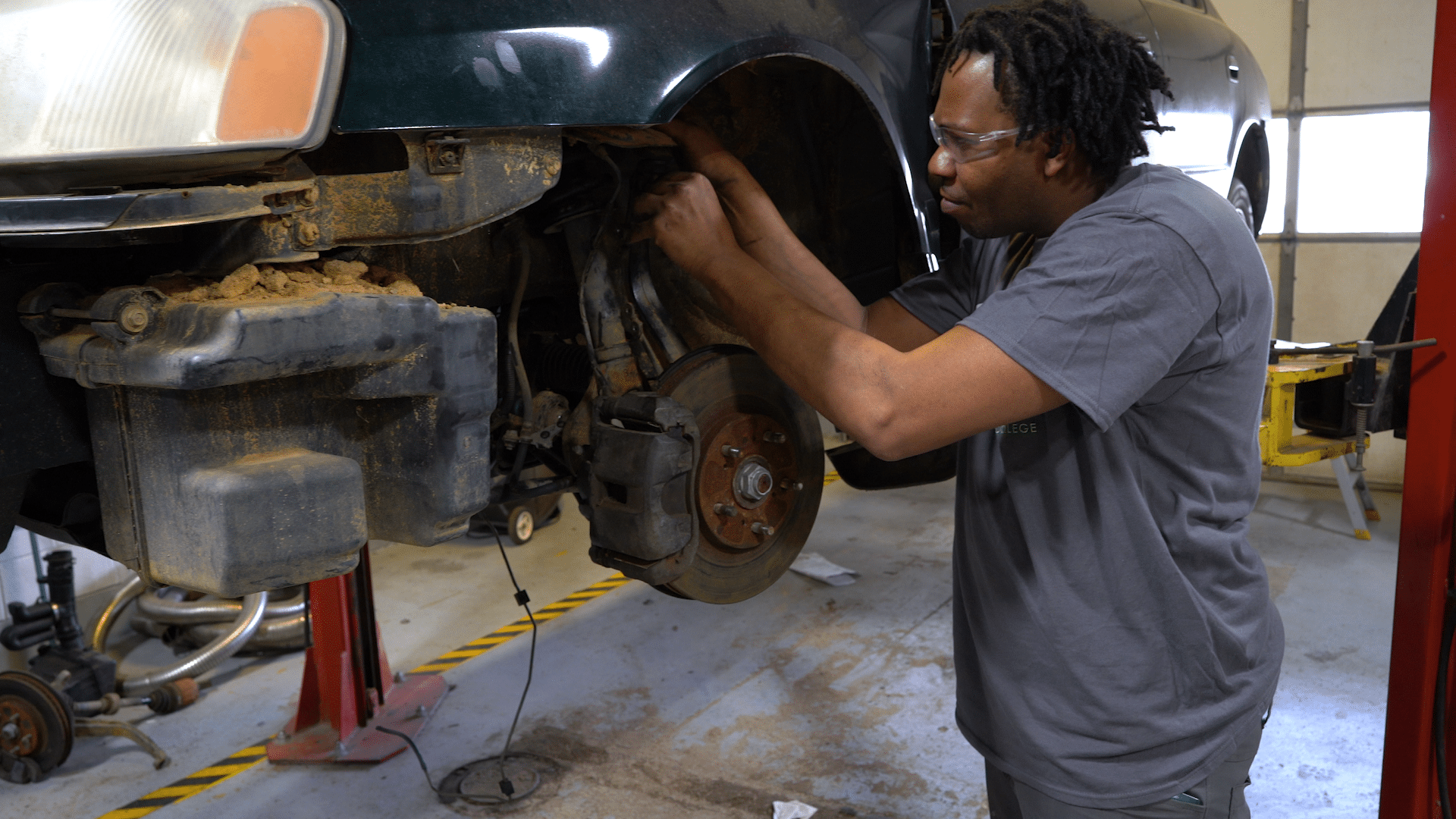 A mechanic wearing safety glasses works on the front wheel area of a car lifted on a hydraulic lift in a garage. The wheel is removed, exposing the brake rotor and suspension components.