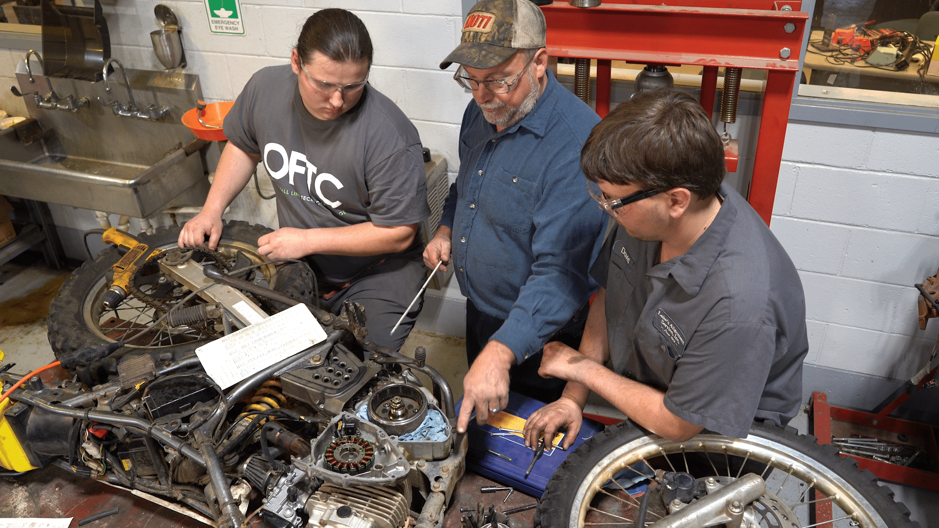 Three men work together on a disassembled motorcycle in a workshop, with tools and parts spread out on the table. One man points to a section of the engine while the others observe closely.