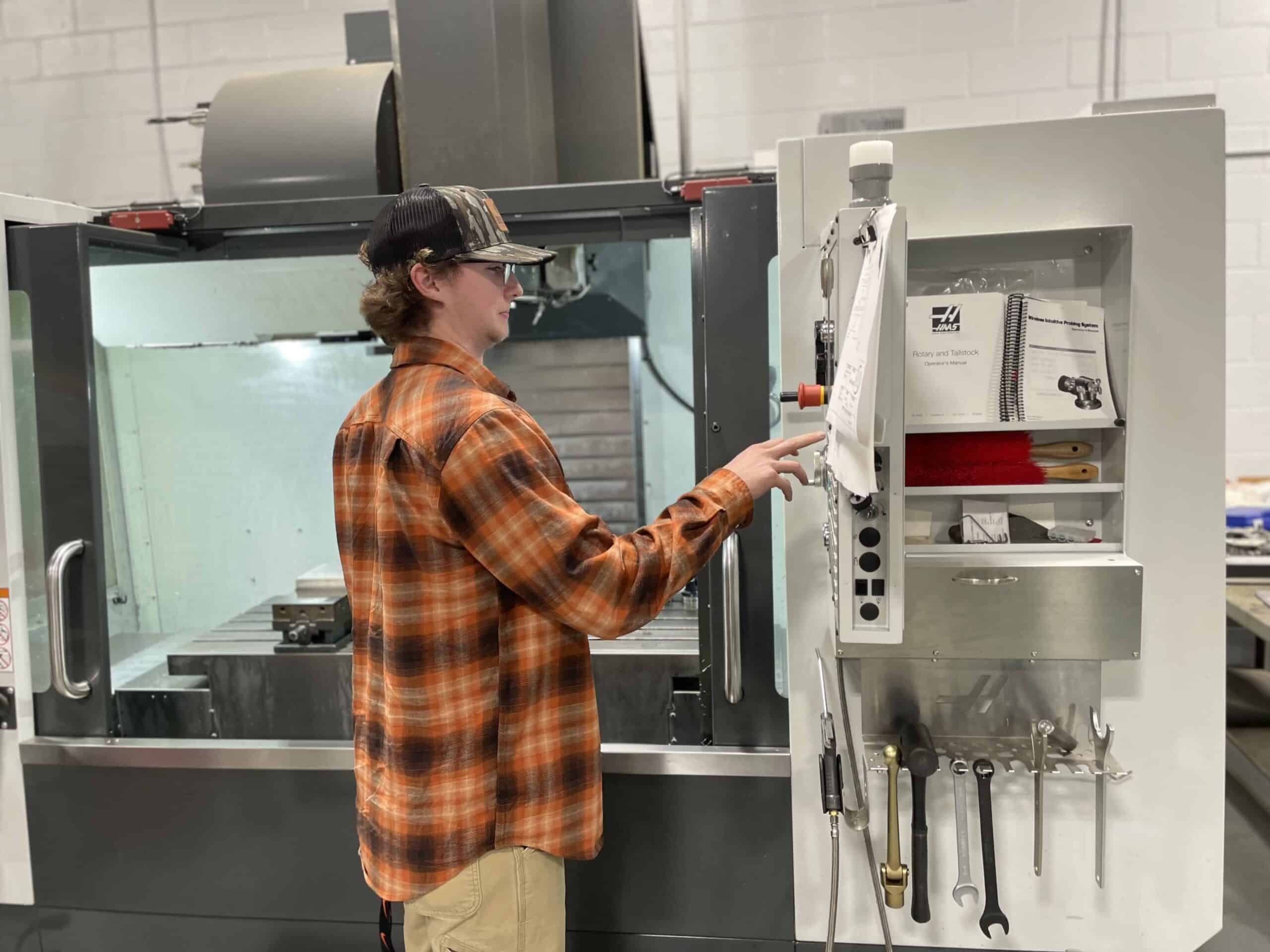 A person in a plaid shirt and cap operates a CNC machine in a workshop, using the control panel. Various tools and manuals are stored on a shelf next to the machine.