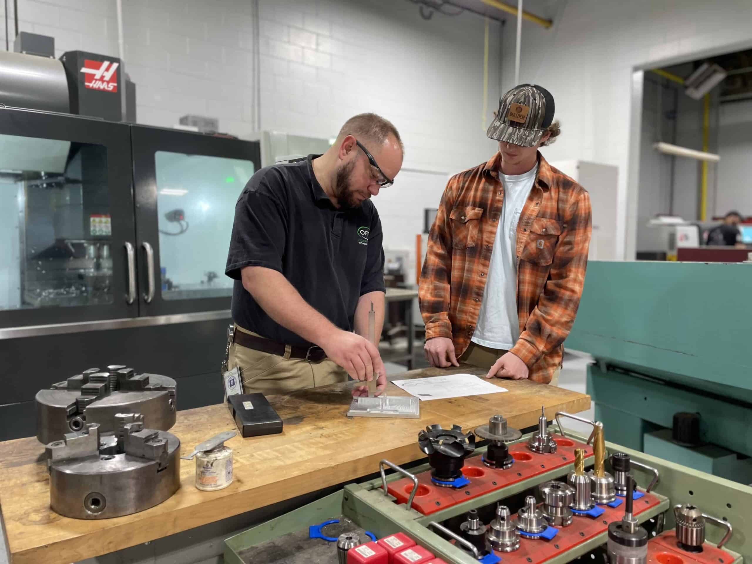 Two men stand at a workbench in a workshop. One, wearing glasses and a black shirt, points to a document while the other, in a plaid shirt and cap, looks on. Metal tools and machinery are visible around them.