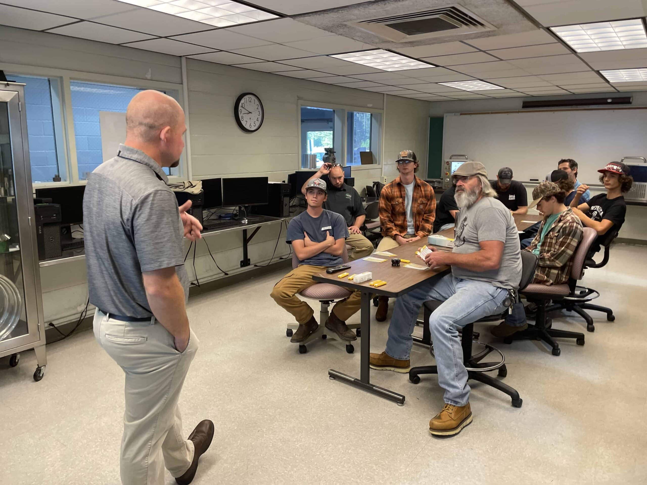 A man stands and speaks to a group of people seated at tables in a classroom. Most listeners wear casual clothes and hats, and some are taking notes. A clock and windows are visible on the wall behind them.