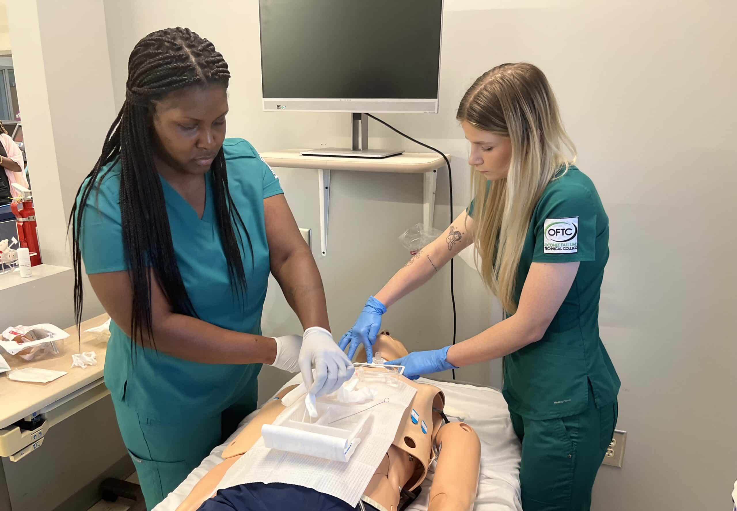 Two medical students in green scrubs practice a medical procedure on a mannequin in a clinical skills lab using gloves and medical supplies with a monitor and equipment nearby.