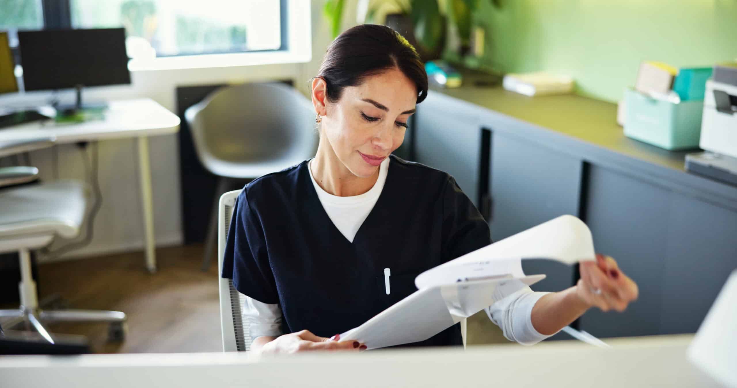 A woman in medical scrubs sits at a desk, looking at papers with a slight smile. She is in a modern office or clinic setting with natural light and plants in the background.