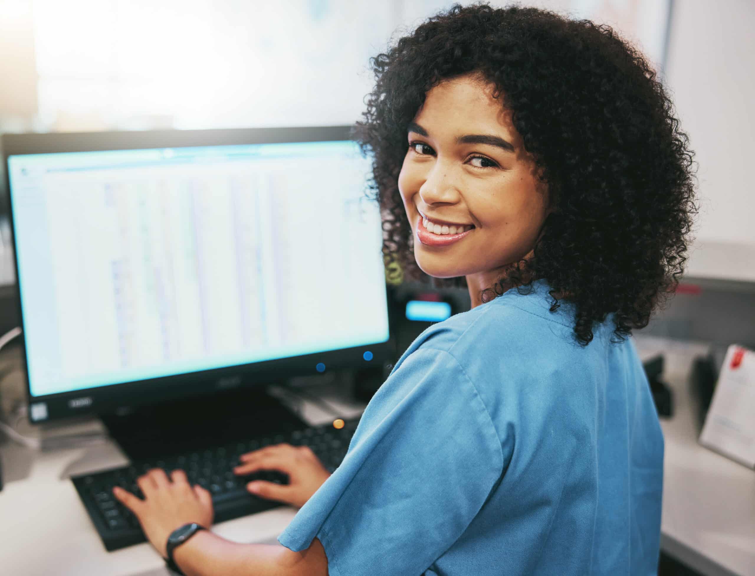 A woman in blue scrubs smiles at the camera while typing on a computer keyboard at a desk, with a large monitor displaying spreadsheets or data in the background.