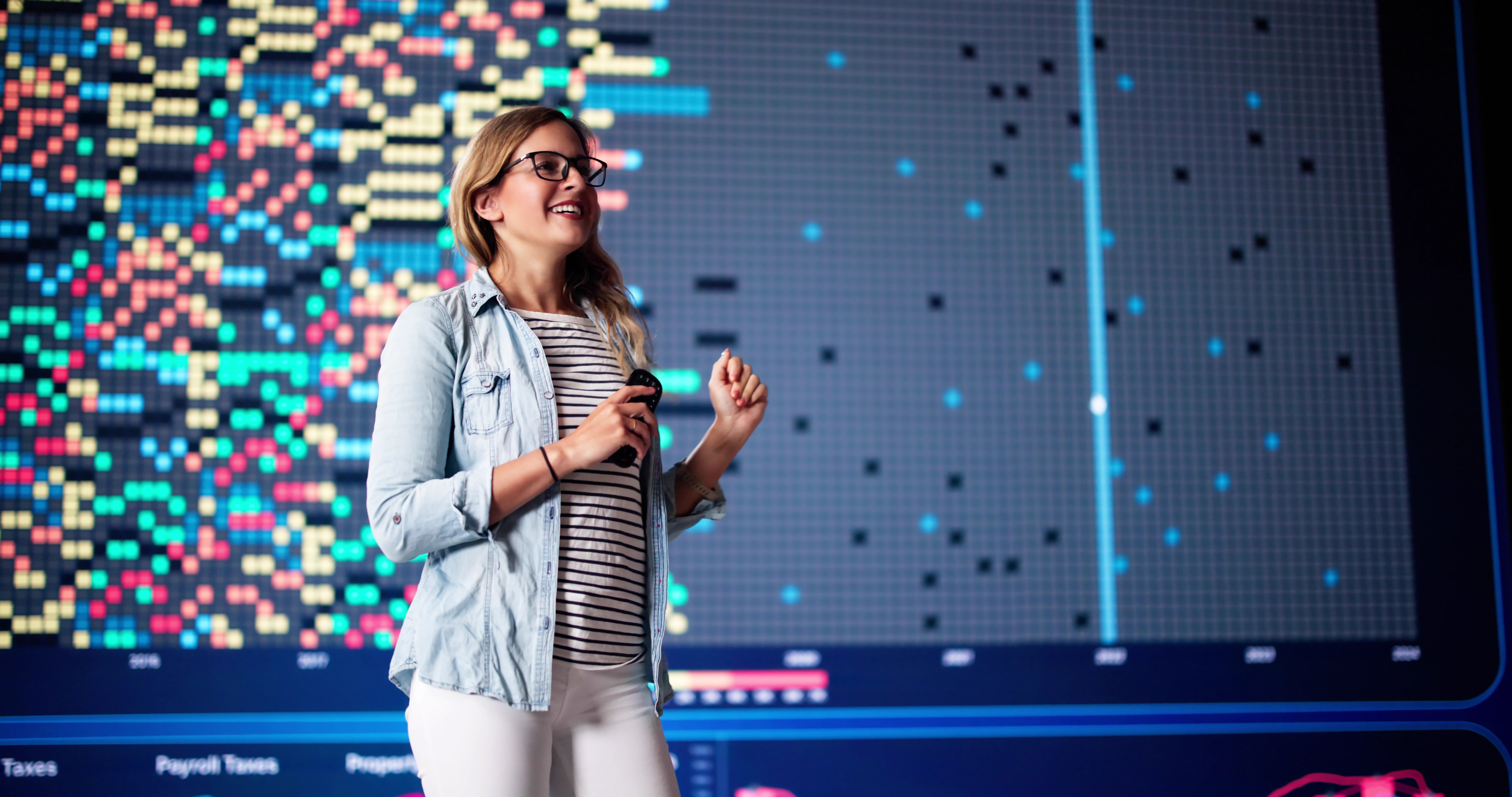 A woman wearing glasses and casual clothes stands smiling in front of a large digital screen displaying colorful charts and data visualizations, holding a microphone or presenter remote.