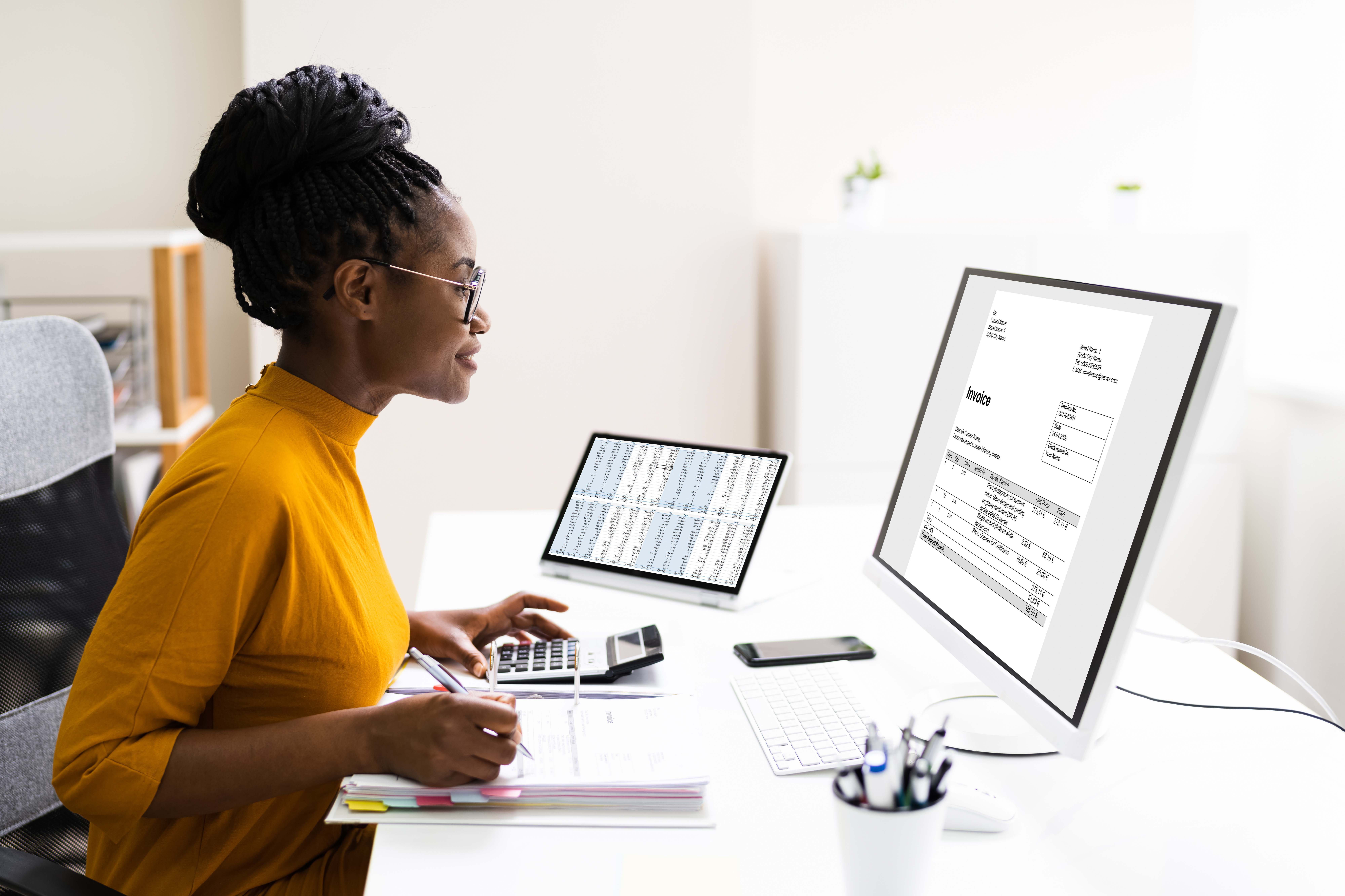 A woman in a yellow top sits at a desk, working on a computer displaying an invoice. She uses a calculator and holds a pen, with financial documents and a laptop showing spreadsheets beside her.
