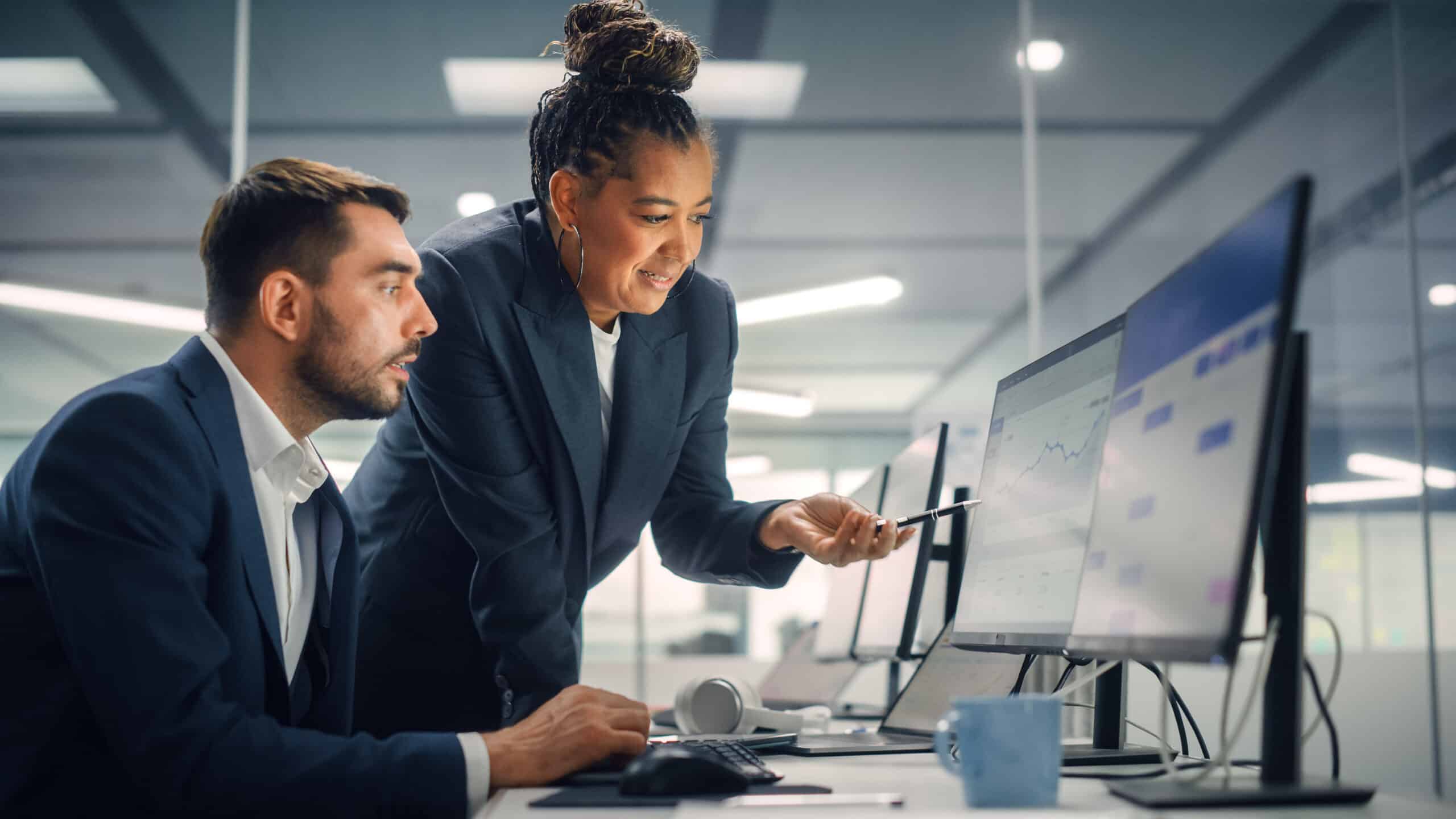 Two business professionals in an office review data charts on a computer screen; one is sitting and typing, while the other stands beside and points at the monitor, both focused on the work.