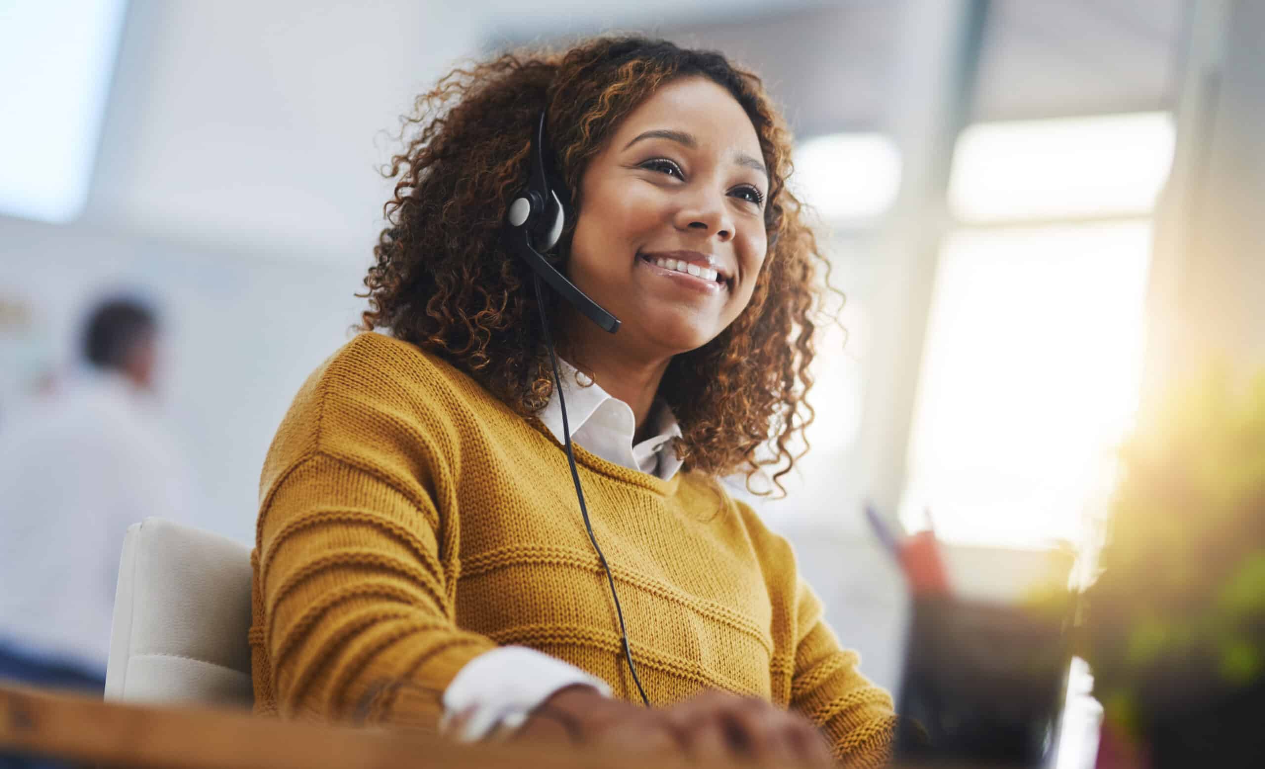 A woman wearing a yellow sweater and a headset smiles while sitting at a desk, suggesting she is working in a customer service or call center environment. Sunlight is visible in the background.