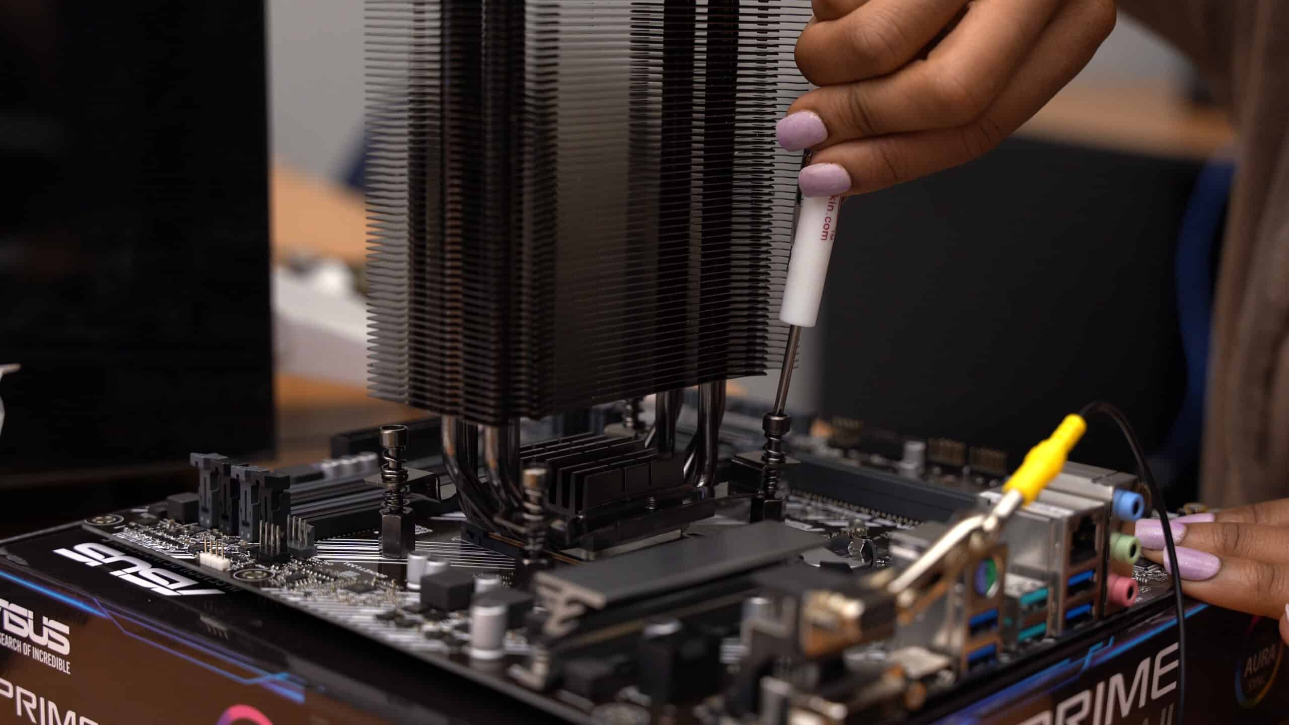A person with light purple nail polish installs a large CPU cooler onto a motherboard, using a screwdriver to secure it. The motherboard rests on top of an ASUS box, with various ports and components visible.