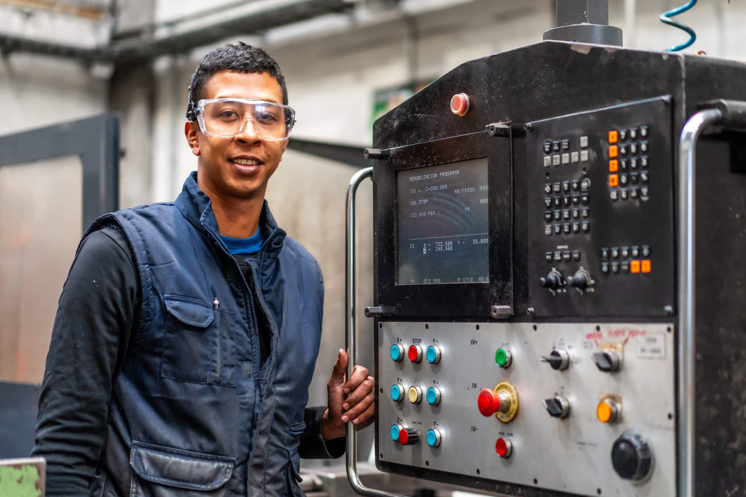 A man wearing safety glasses and a navy vest stands beside an industrial control panel with various buttons and a display screen, smiling at the camera in a factory setting.
