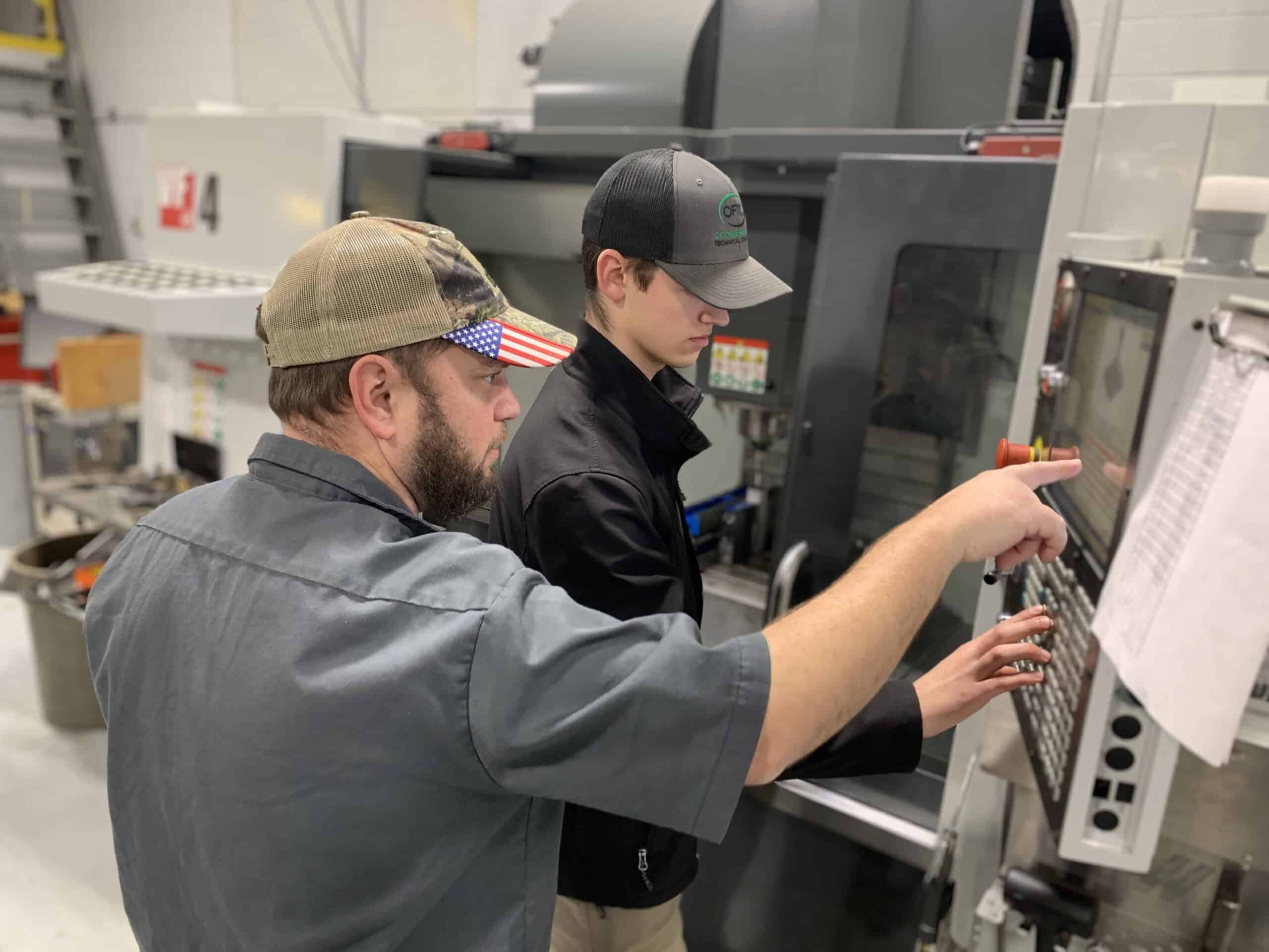 Two men, one older and one younger, operate a CNC machine. The older man, wearing a cap with a US flag, points to the control panel while instructing the younger man, who is also wearing a cap.