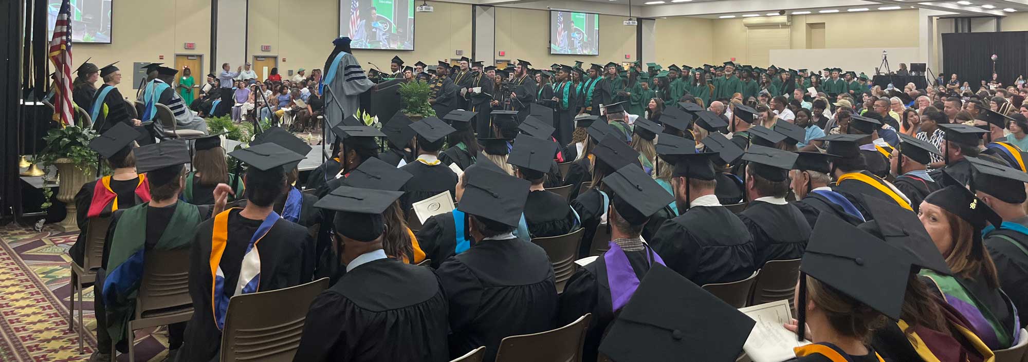 A large group of graduates in caps and gowns sit facing a stage during an indoor graduation ceremony, with an audience of family and friends seated in the background.