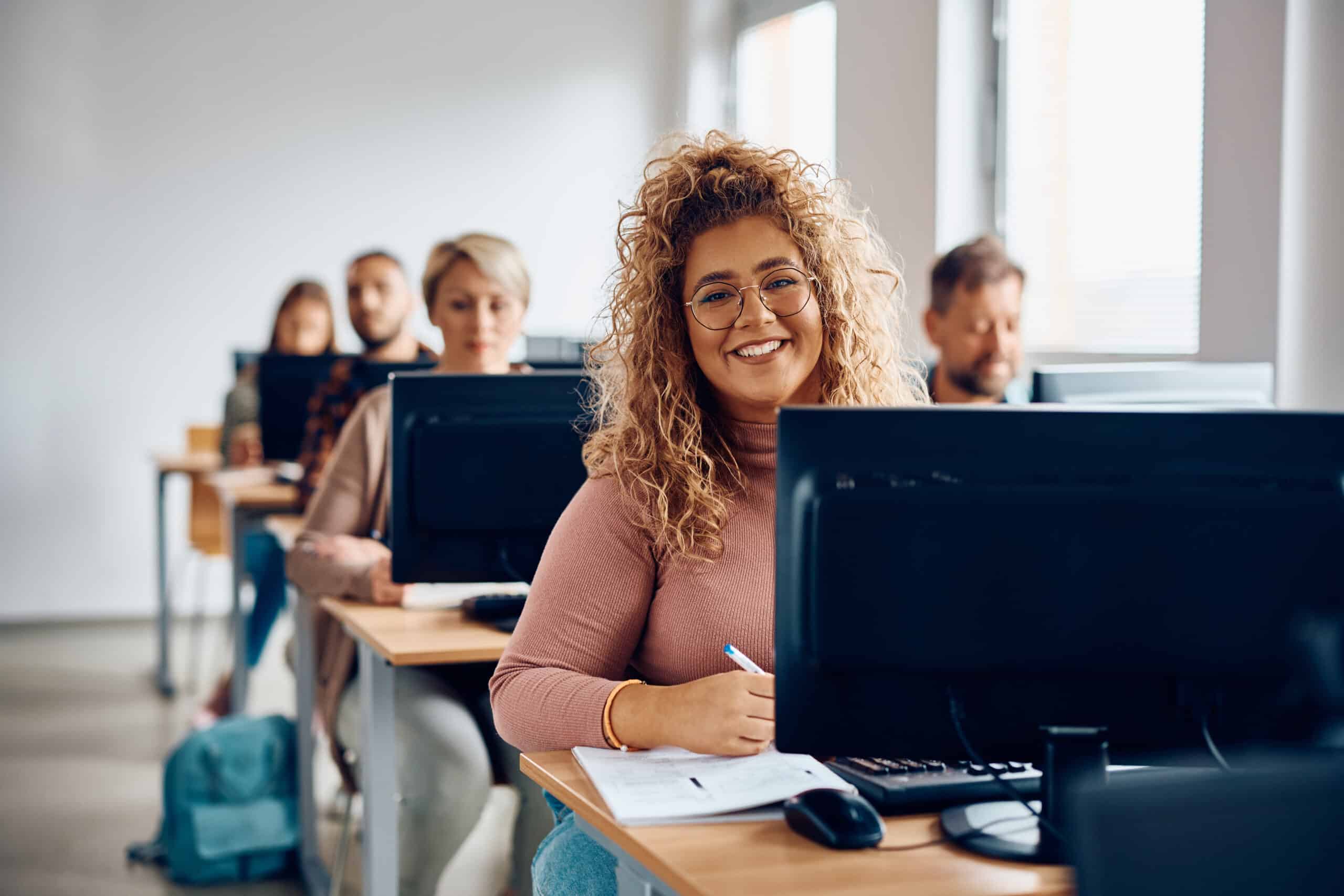 A young woman with curly hair and glasses smiles at the camera while sitting at a desk in a computer classroom, surrounded by other students working on desktop computers.