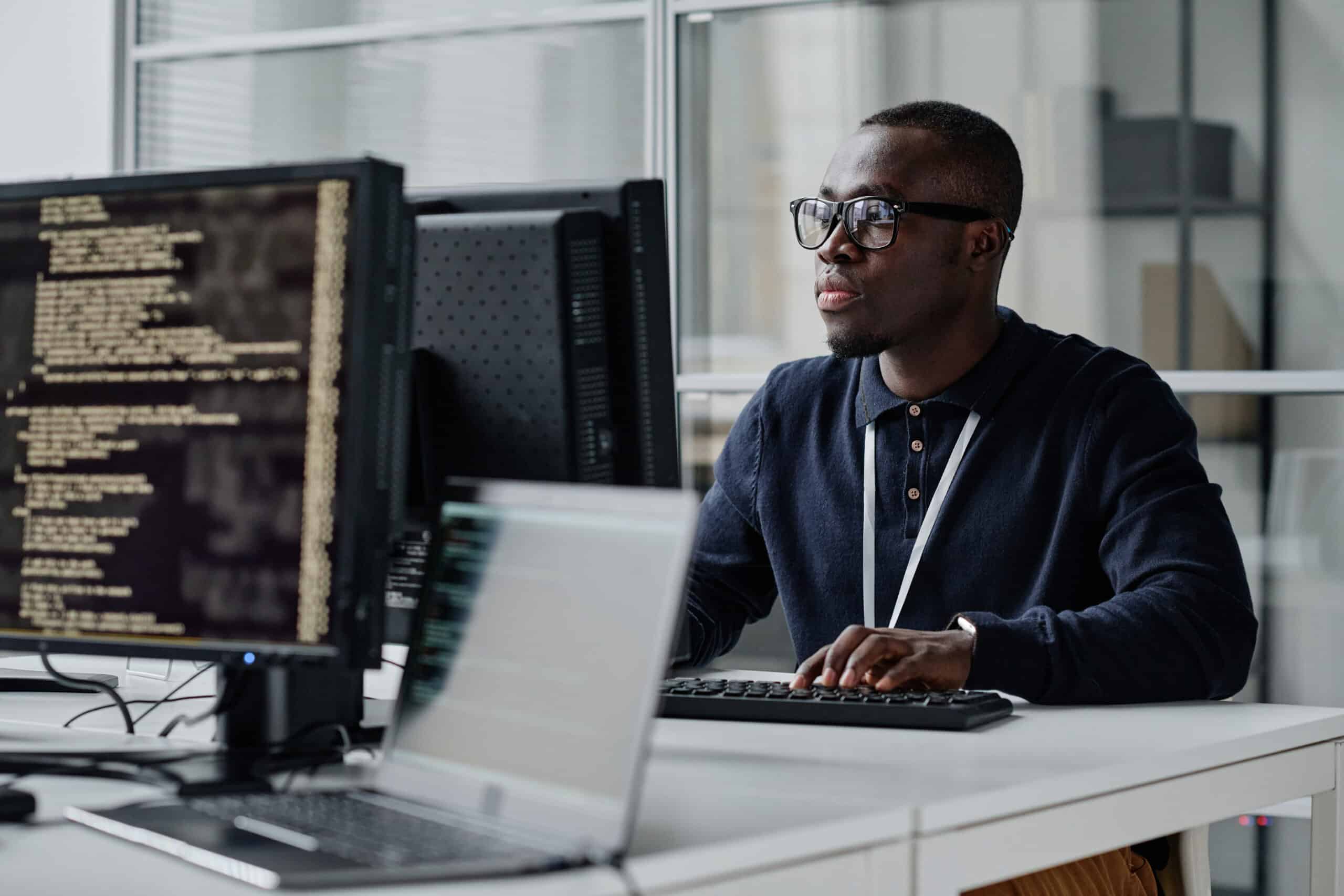 A man wearing glasses works at a desk with computer monitors displaying code in a modern office environment. He is focused on the screen and typing on a keyboard.