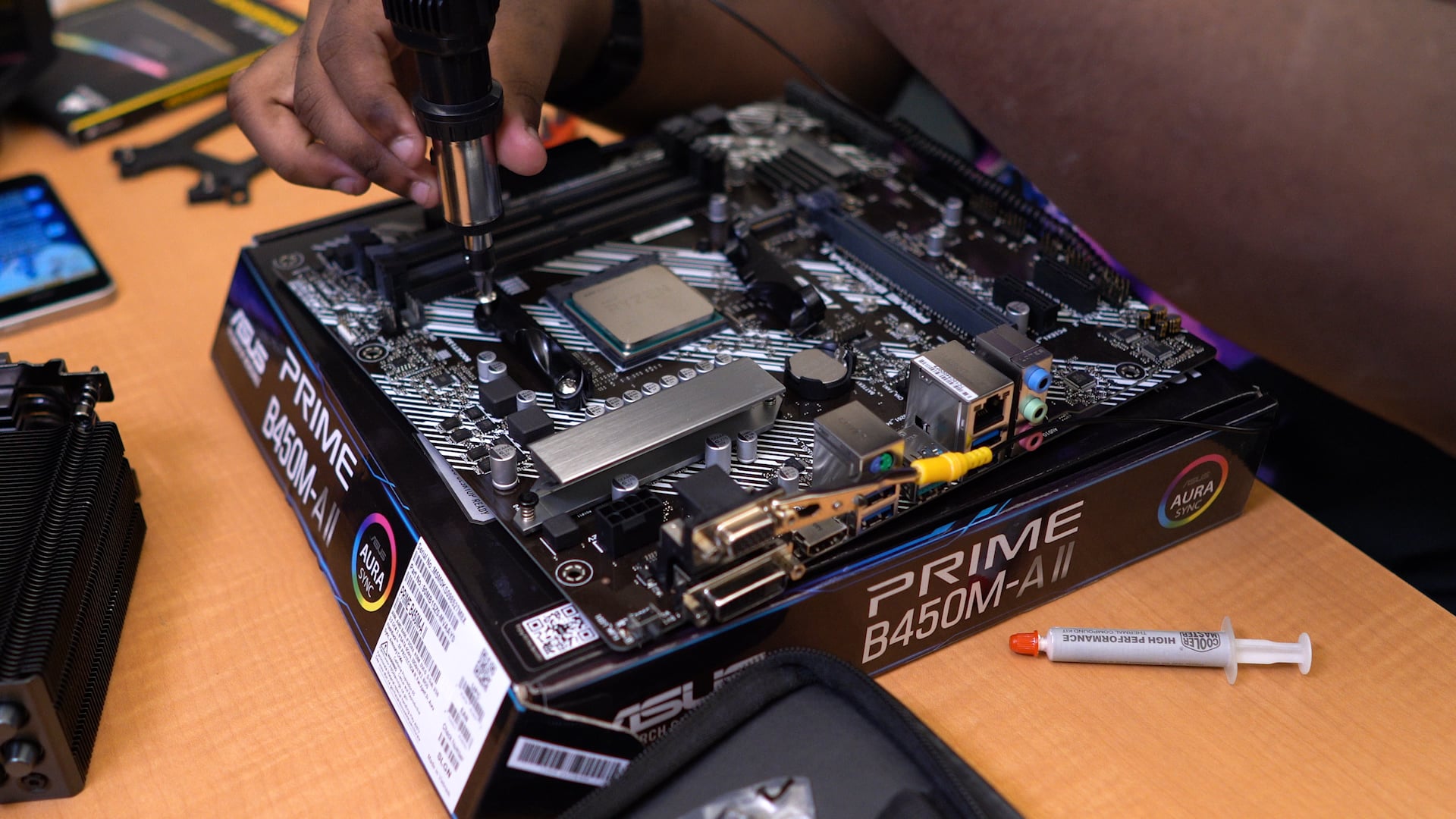 A person uses a power tool to assemble a computer motherboard on top of an ASUS PRIME B450M-A/II box; a syringe of thermal paste and computer parts are nearby on the table.
